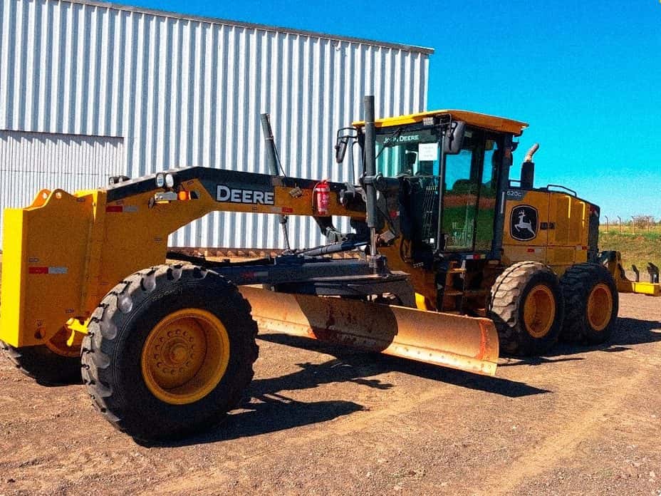 Motoniveladora John Deere 620G com lâmina central, ao lado do galpão da empresa